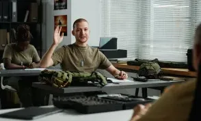 Young male student raising hand and looking at teacher at lesson while asking him question after lecture about military training